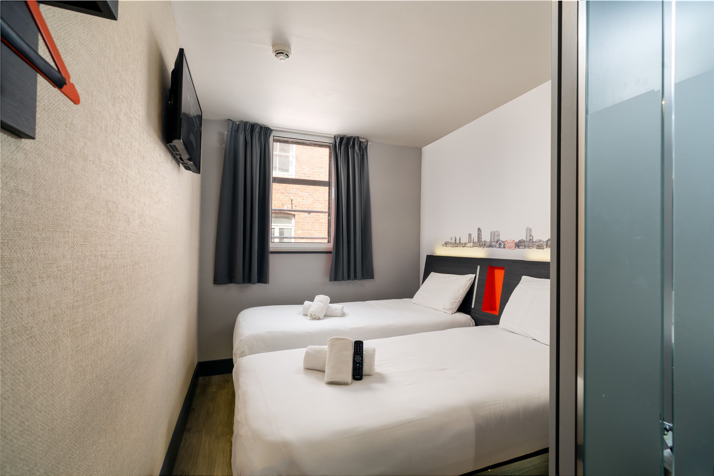 easyHotel Triple Room in Glasgow, featuring two single beds with crisp white bedding, a wall-mounted television, and a window with dark curtains. A distinctive city skyline graphic is displayed above the headboards.
