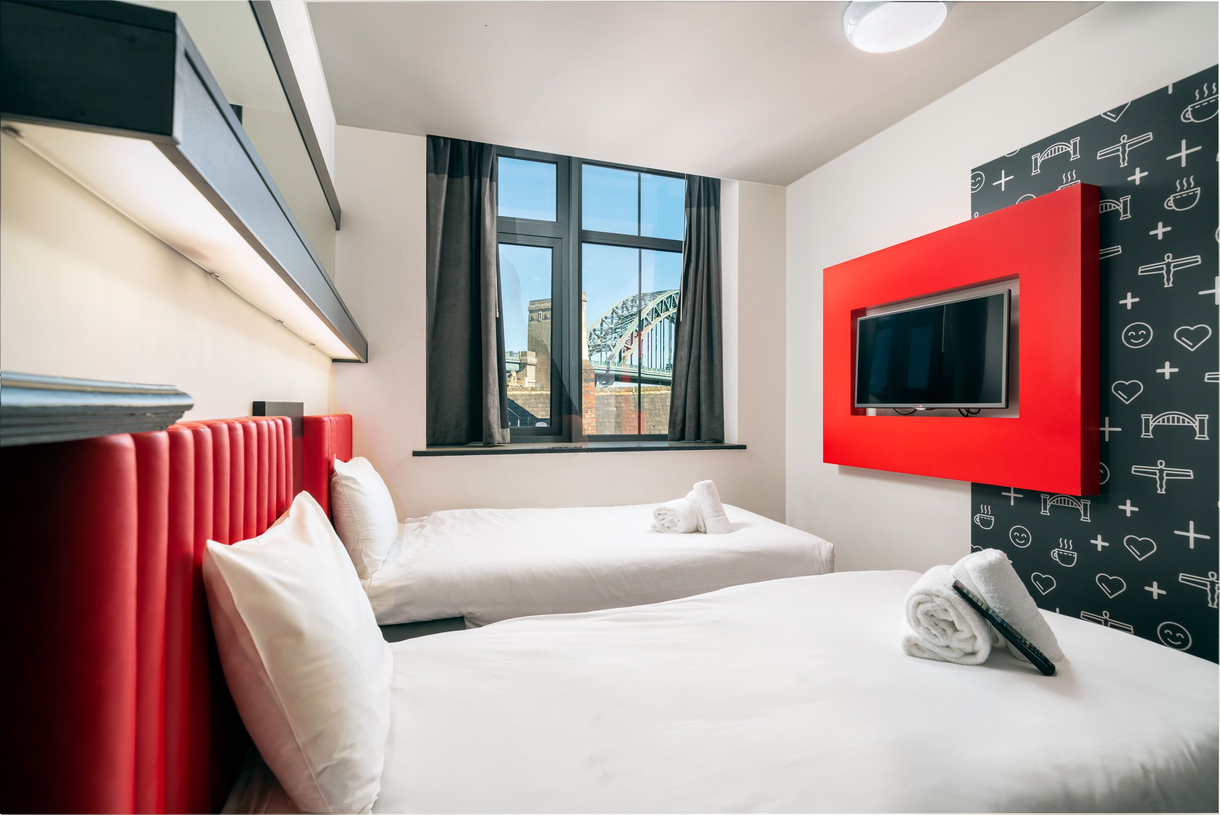 An easyHotel twin room in Newcastle upon Tyne, UK, featuring two beds with red headboards, a wall-mounted TV, and a clear view of the Tyne Bridge from the window.