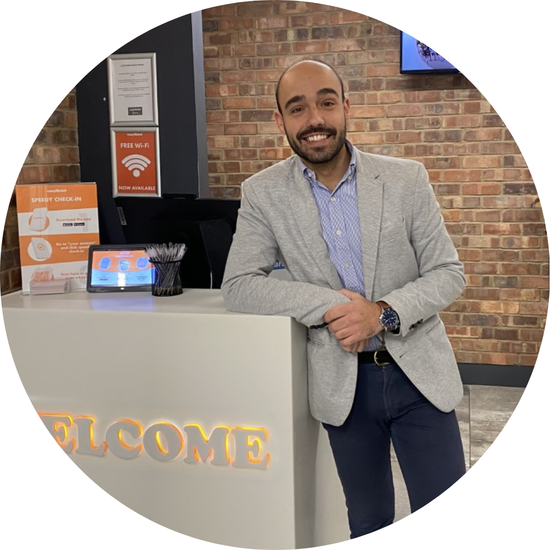 Smiling easyHotel staff member at the hotel reception desk, with a 'WELCOME' sign, 'Speedy Check-in', and 'Free Wi-Fi' signage in the background.