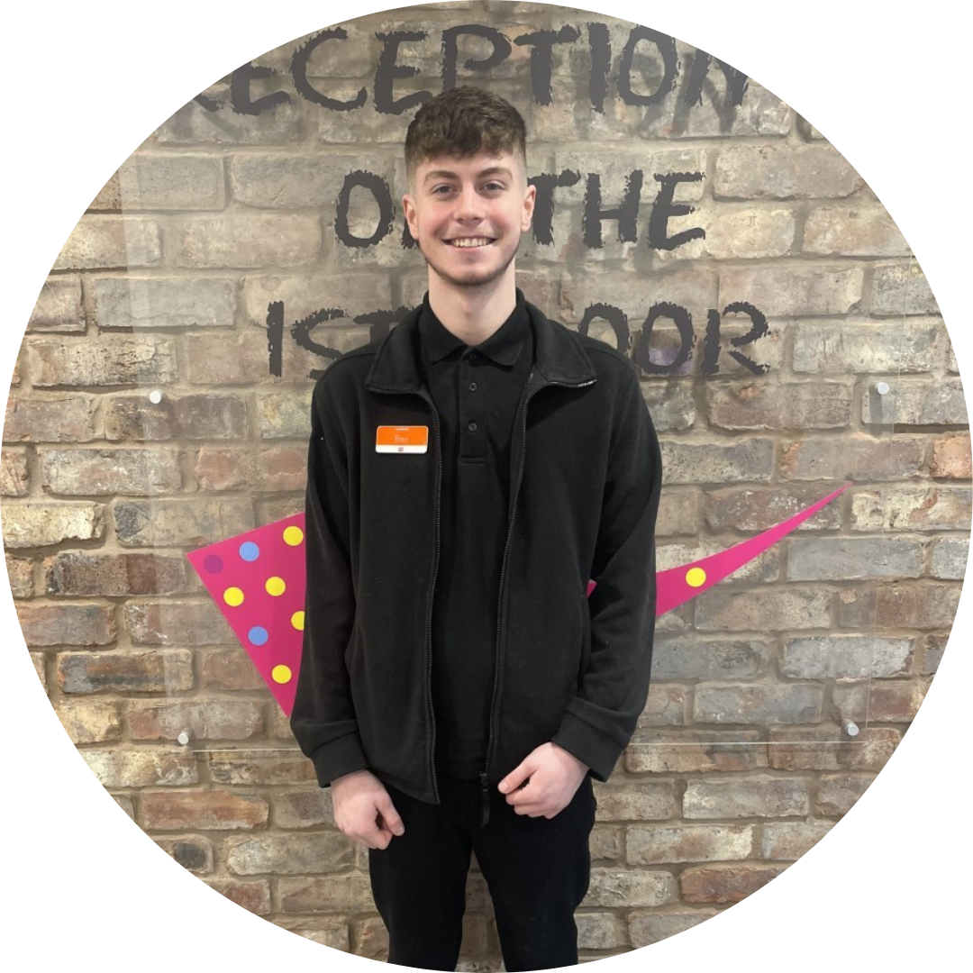 Smiling easyHotel staff member Ben in a hotel lobby, with 'Reception on the 1st floor' signage on a brick wall.
