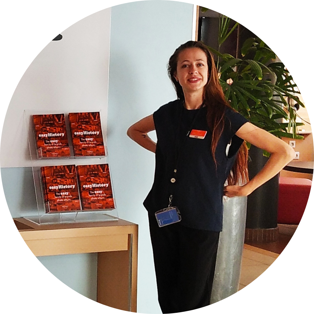 Smiling easyHotel staff member in a modern hotel lobby next to a display of easyHistory brand photo albums.