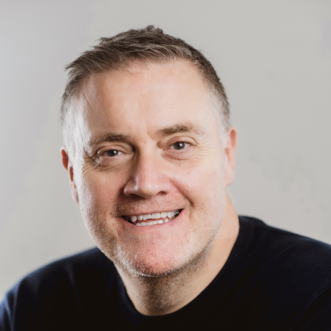 Headshot of a smiling middle-aged man with short, greying hair, wearing a dark shirt against a plain light background.