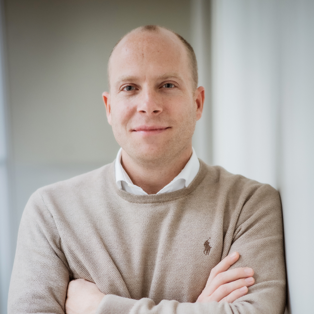 Professional portrait of a smiling man in a beige sweater with crossed arms.