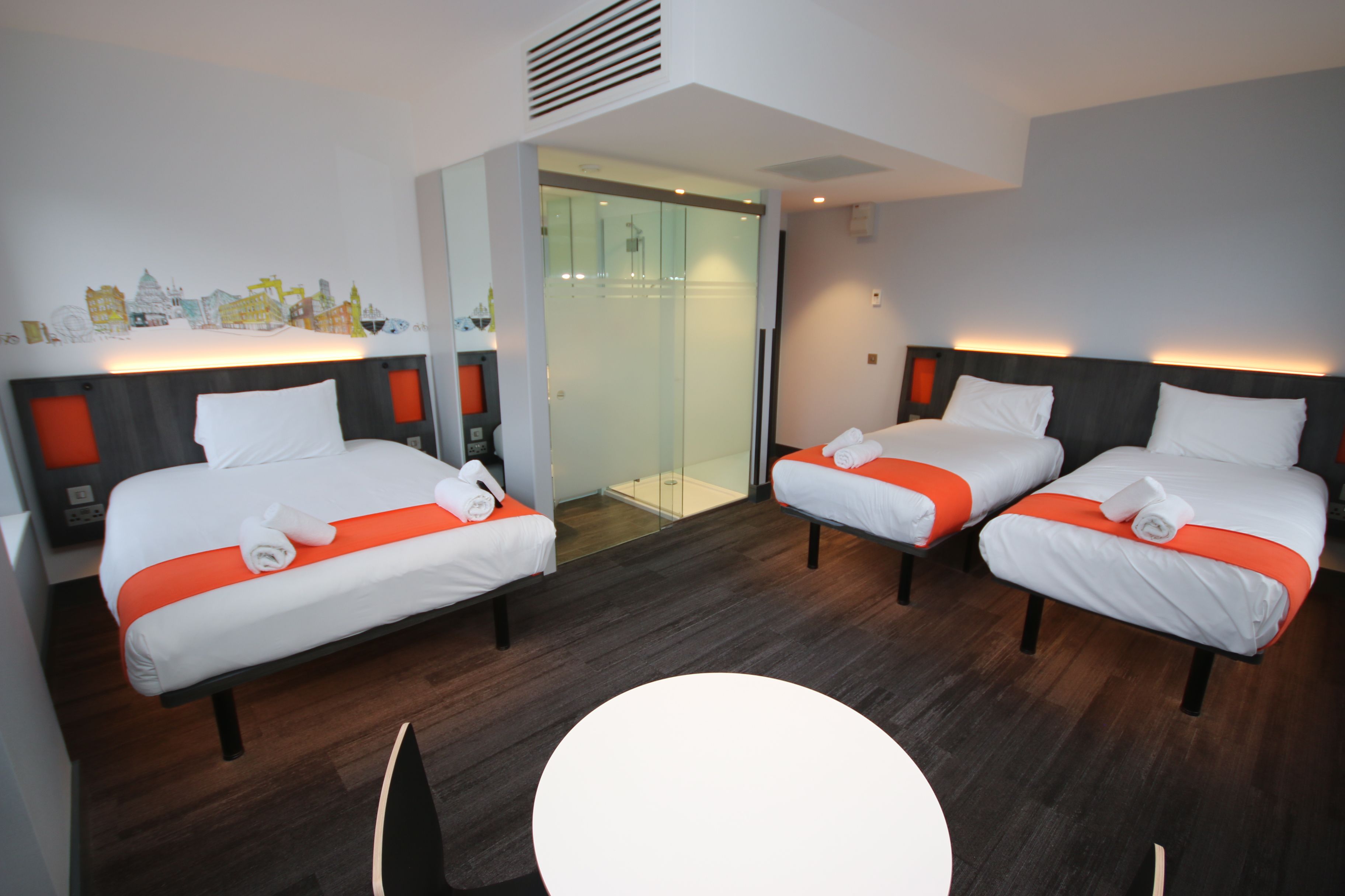 A bright, modern easyHotel family room in Belfast with three beds featuring white linen and orange accents, rolled towels, a cityscape mural, and an open-plan glass shower.