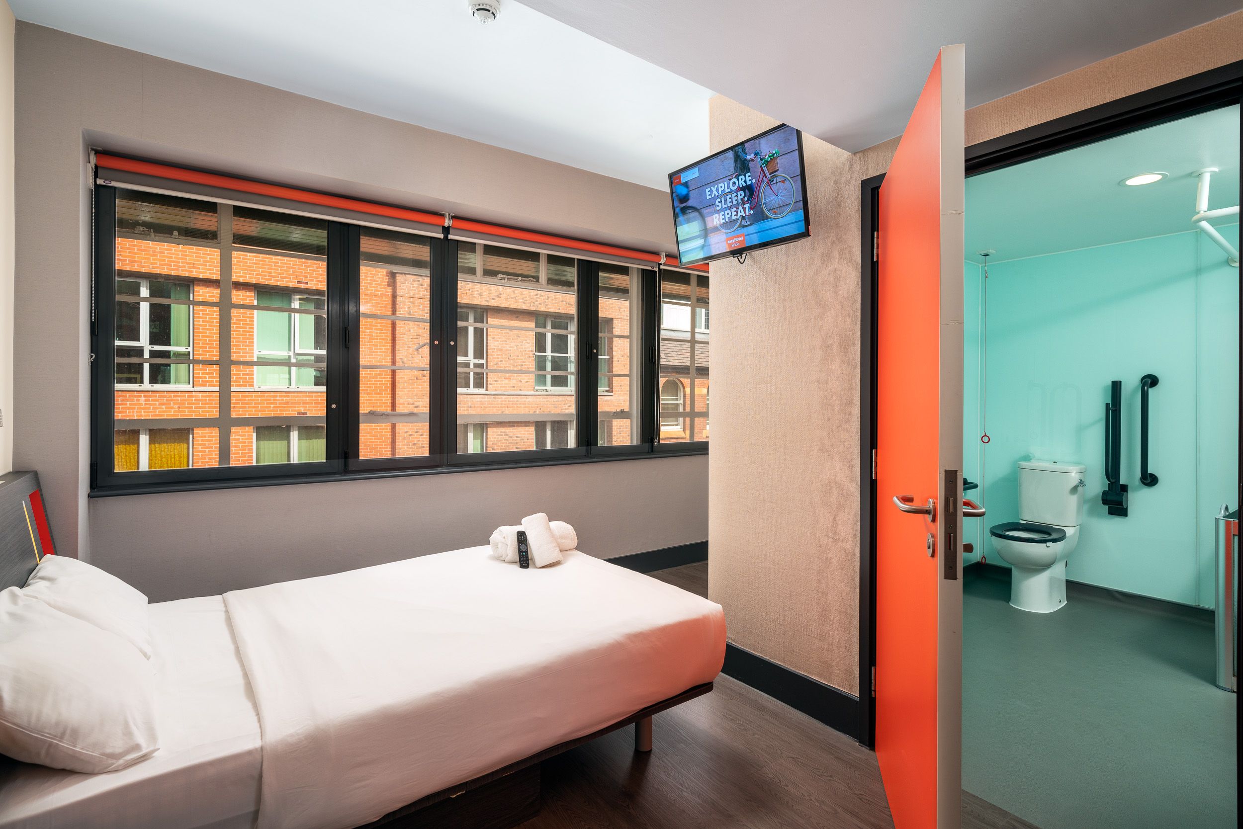 Accessible easyHotel room in Birmingham with a single bed, wall-mounted TV, and an orange door leading to a teal accessible bathroom with grab bars. A large window offers a view of urban brick buildings.