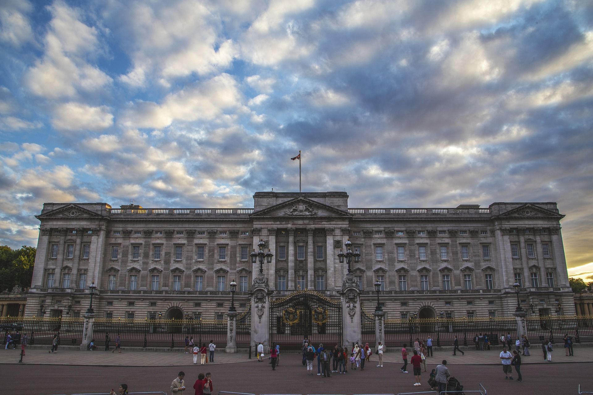 Buckingham Palace in London, a grand stone building with the British flag flying, under a dramatic cloudy sky, with many visitors gathered in front of its ornate gates.