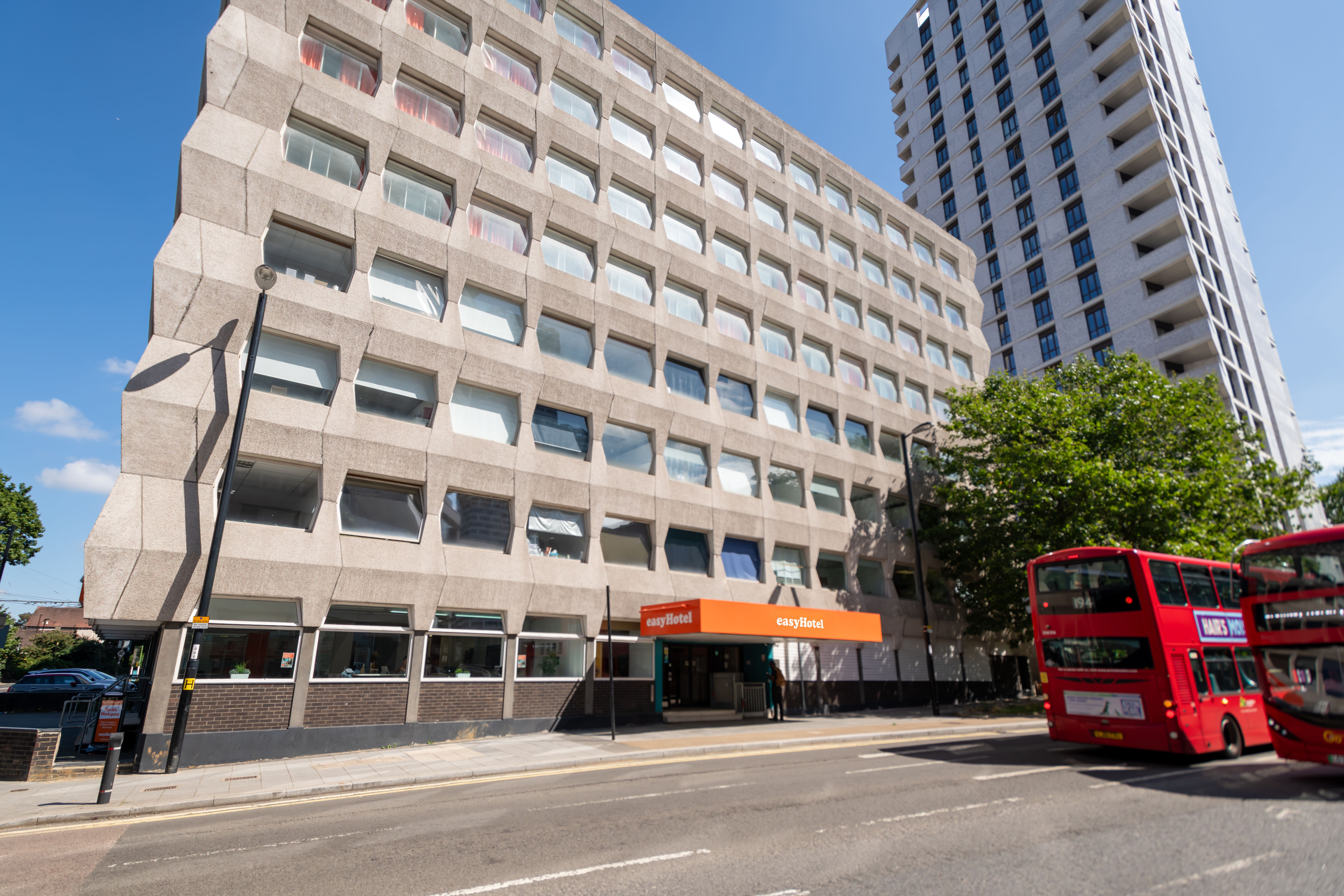 Exterior of easyHotel Croydon, a large brutalist building with an angular concrete facade and a prominent orange entrance canopy, on a sunny day with red double-decker buses on the street.