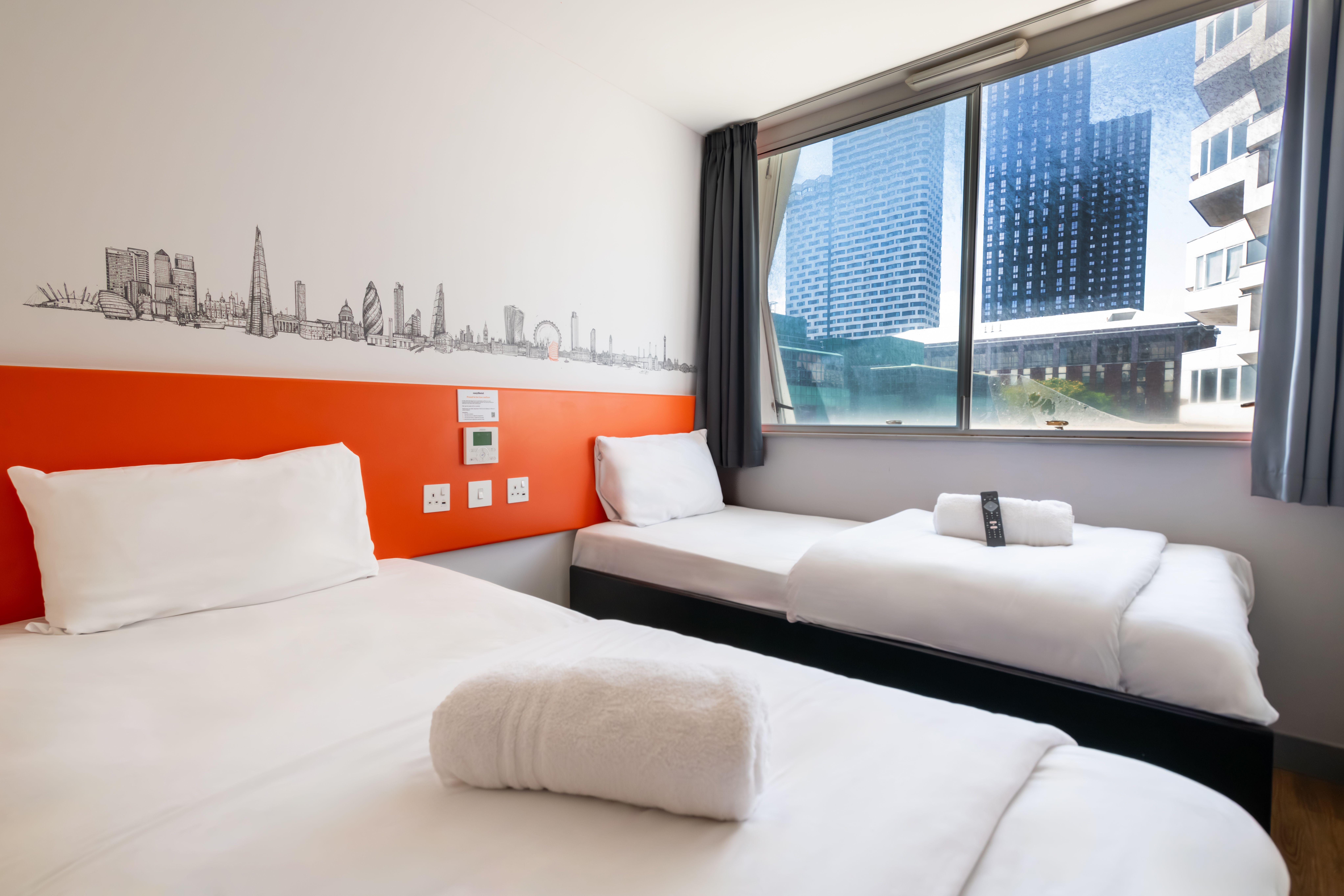 An easyHotel twin room in Croydon featuring two single beds with white bedding, an orange headboard with UK power outlets, and a black and white London skyline mural. A large window offers a view of tall city buildings.