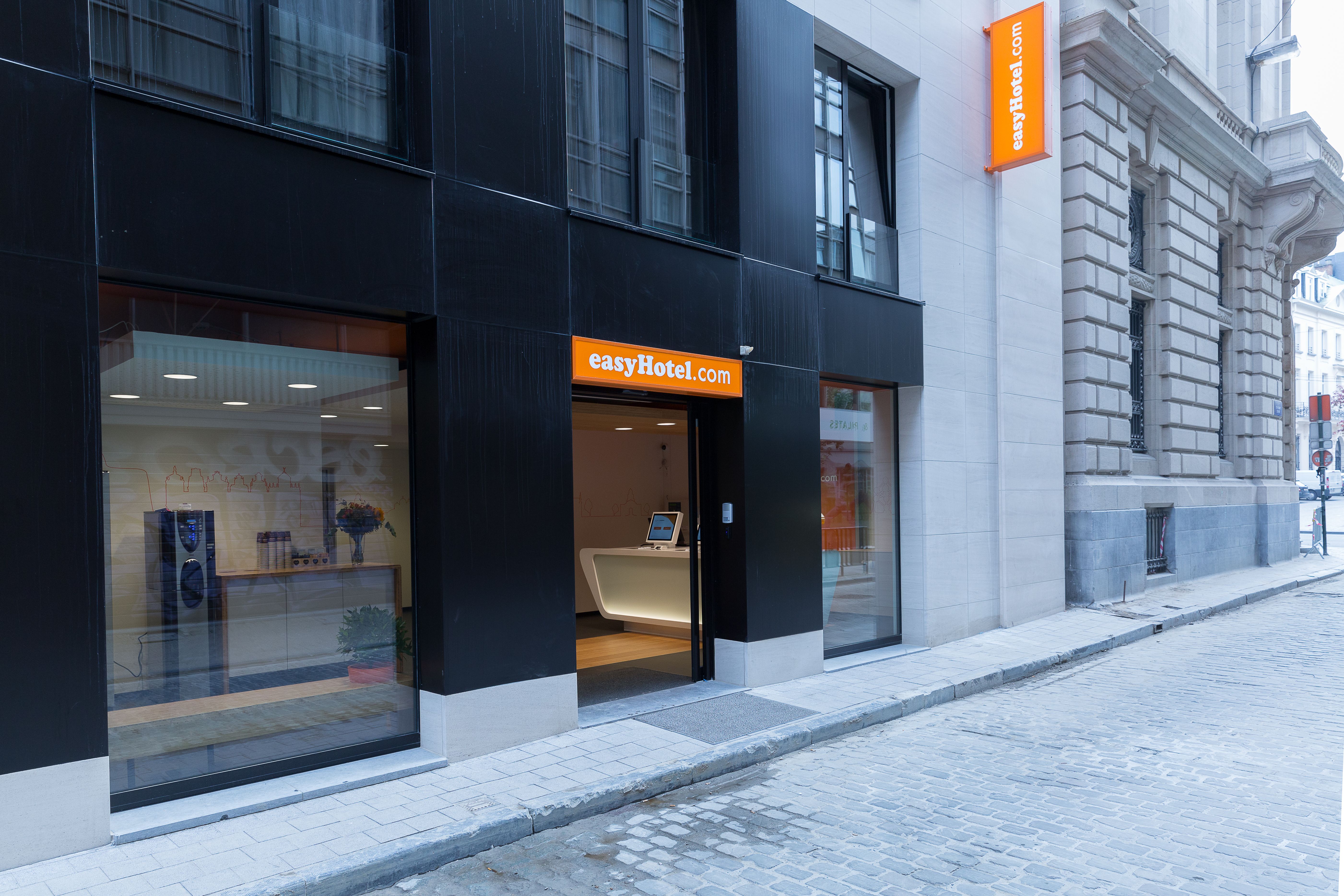 A street-level view of the easyHotel Brussels entrance, featuring a sleek black facade with prominent orange easyHotel.com signage, a visible modern lobby with a self-service check-in counter, and a wet cobblestone street.