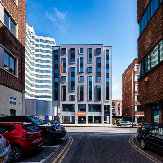 Exterior view of the modern easyHotel Cardiff building under a blue sky. The grey facade features an irregular pattern of large and small rectangular windows with orange and brown frames. Parked cars are visible on the street in the foreground, with other