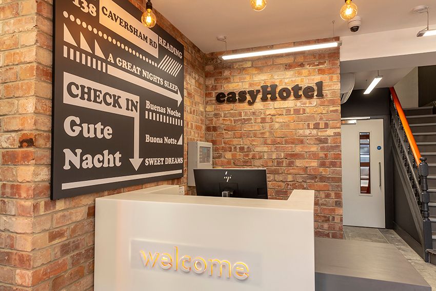 easyHotel Reading hotel lobby featuring a modern white reception desk with an illuminated "welcome" sign, exposed brick walls, and a large black and white sign displaying the address at 138 Caversham Road and hotel information. The easyHotel logo is promi