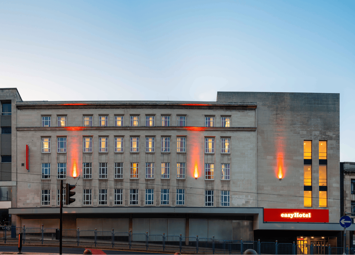 Exterior view of the easyHotel Sheffield building at dusk, featuring its stone facade illuminated by orange uplights and glowing windows, with a prominent red easyHotel sign at street level.