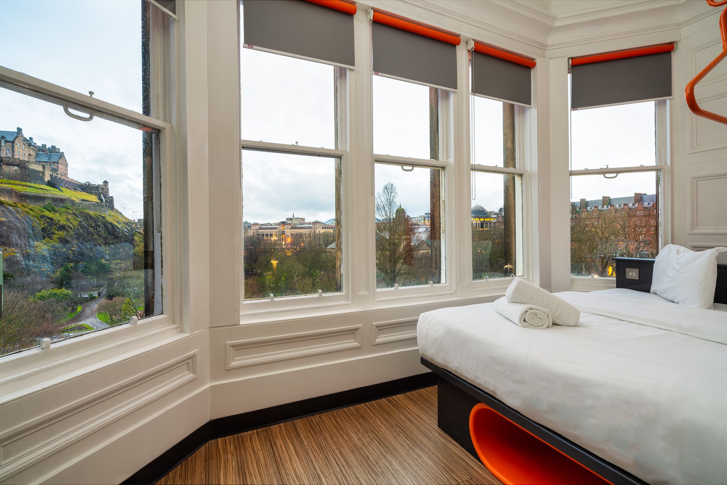 An easyHotel room in Edinburgh with a comfortable bed, white linen, and stacked towels, featuring large bay windows offering a direct view of Edinburgh Castle and the city's cityscape.