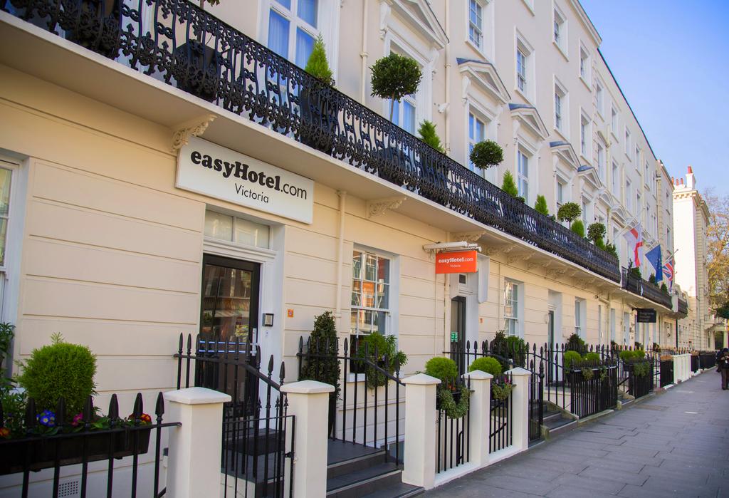 Exterior view of the easyHotel Victoria in London, featuring a cream-colored facade with black wrought-iron balconies, manicured plants, and flags.