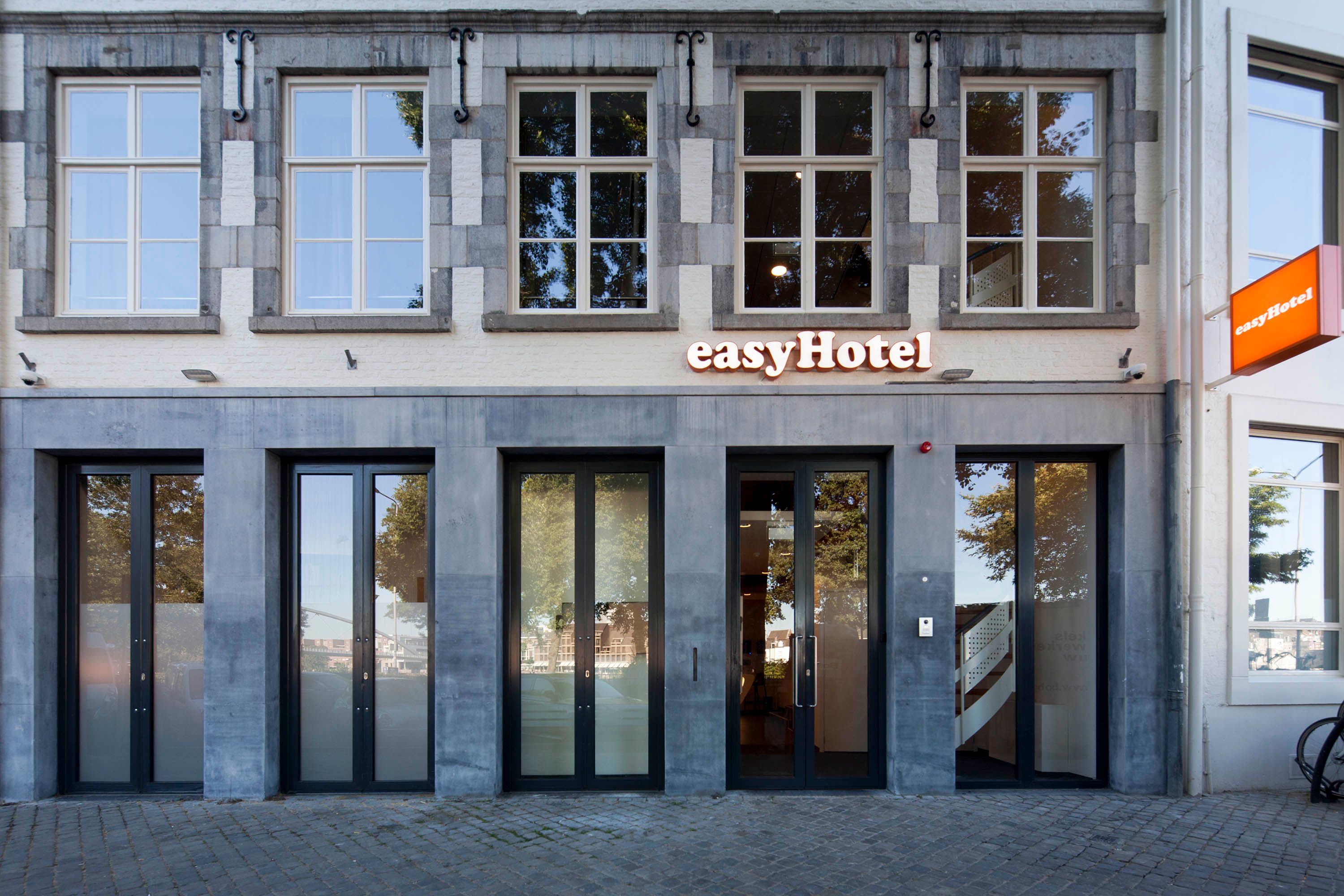 Exterior facade of easyHotel Maastricht, featuring a distinctive gray stone and white brick building with multiple large windows and glass entrance doors on a cobblestone street.