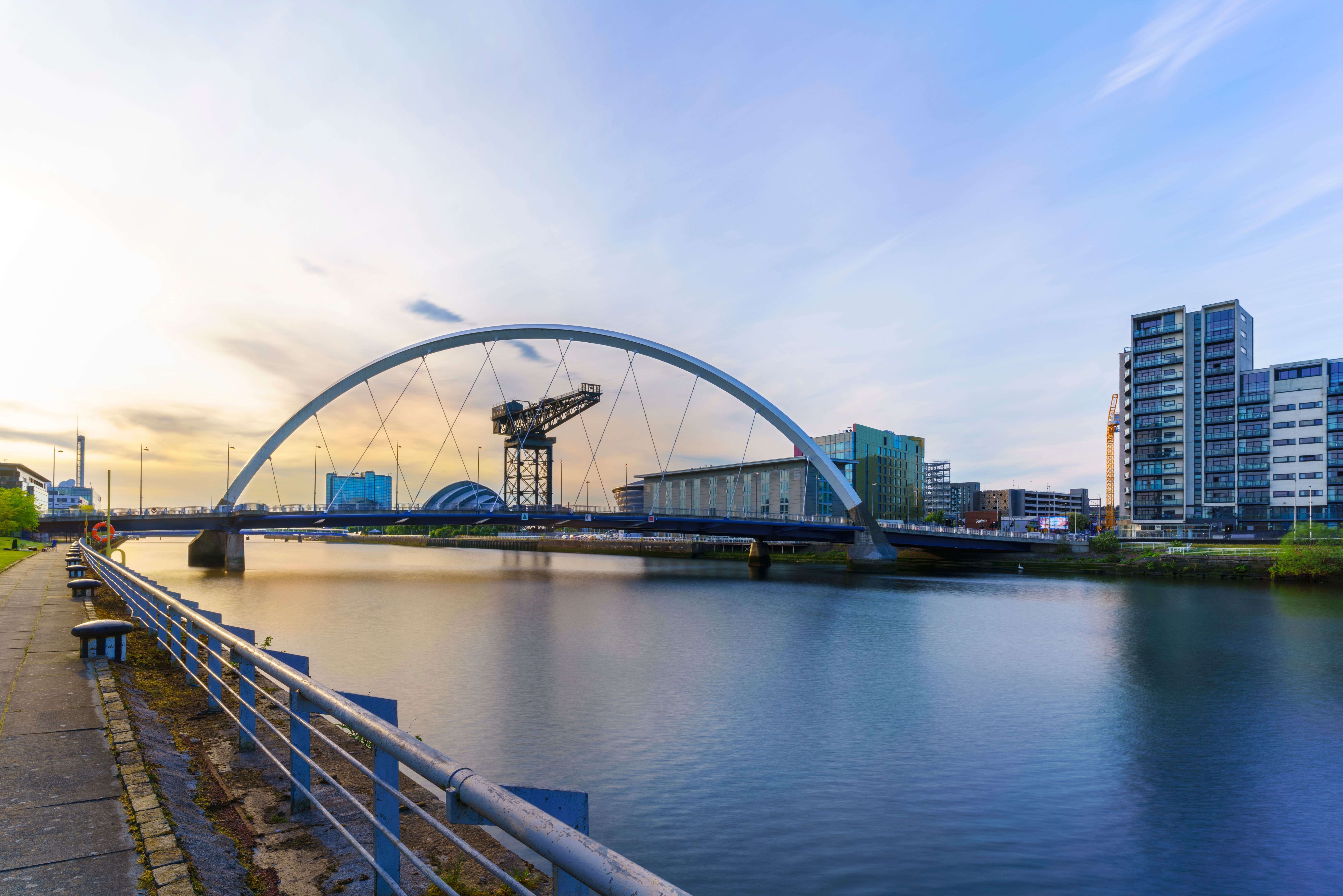 Evening view of Glasgow's River Clyde, featuring the iconic Clyde Arc bridge, Finnieston Crane, and SEC Armadillo, with modern city buildings under a colorful sky.