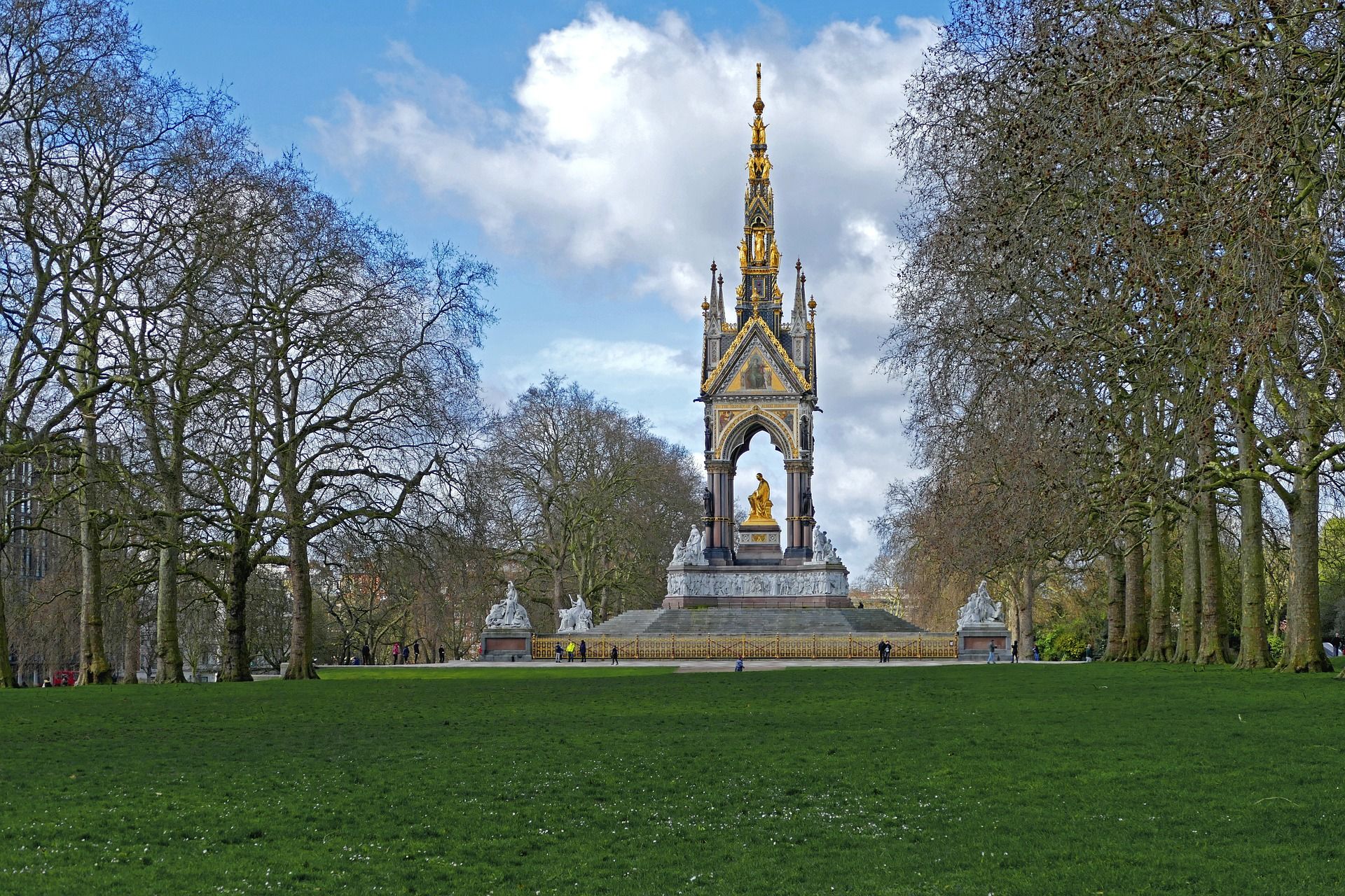 The grand Albert Memorial, a golden Gothic monument, stands amidst green lawns and trees in Kensington Gardens, London, near Hyde Park, under a partly cloudy sky.