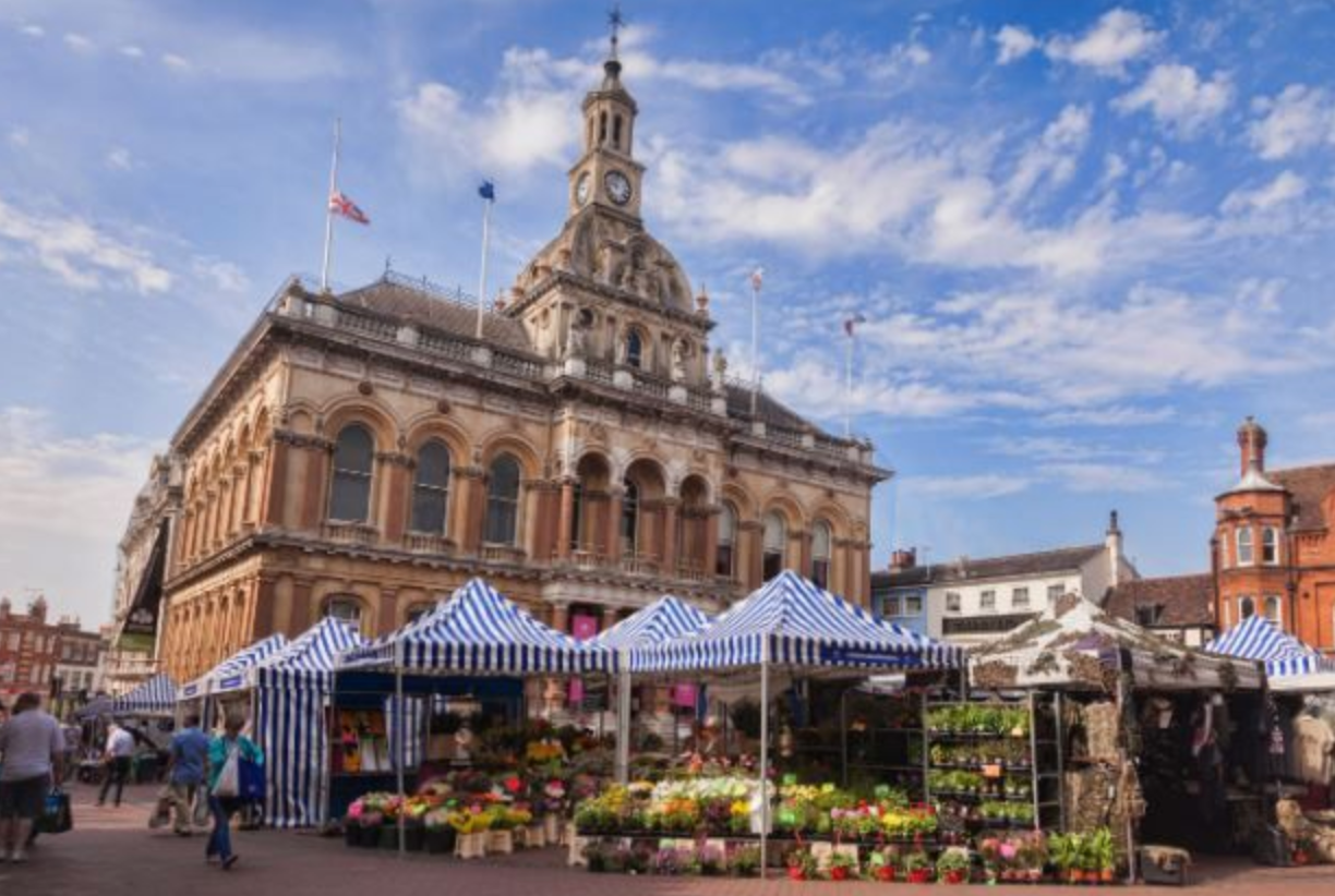 A bustling market scene in front of the historic Ipswich Corn Exchange building, featuring blue and white striped stalls filled with colorful flowers and plants in the town center.