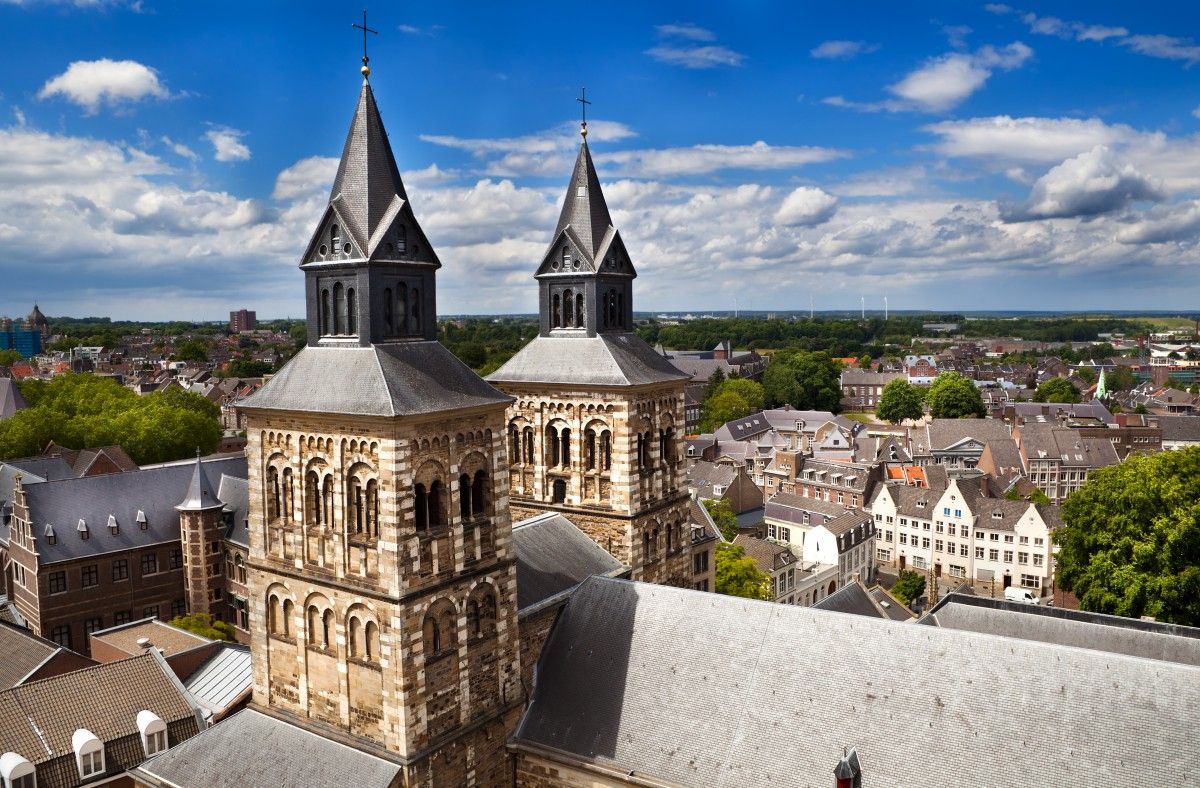 Panoramic view of Maastricht, Netherlands, showcasing two historic stone church towers with spires rising above traditional city rooftops under a bright blue sky with scattered clouds.