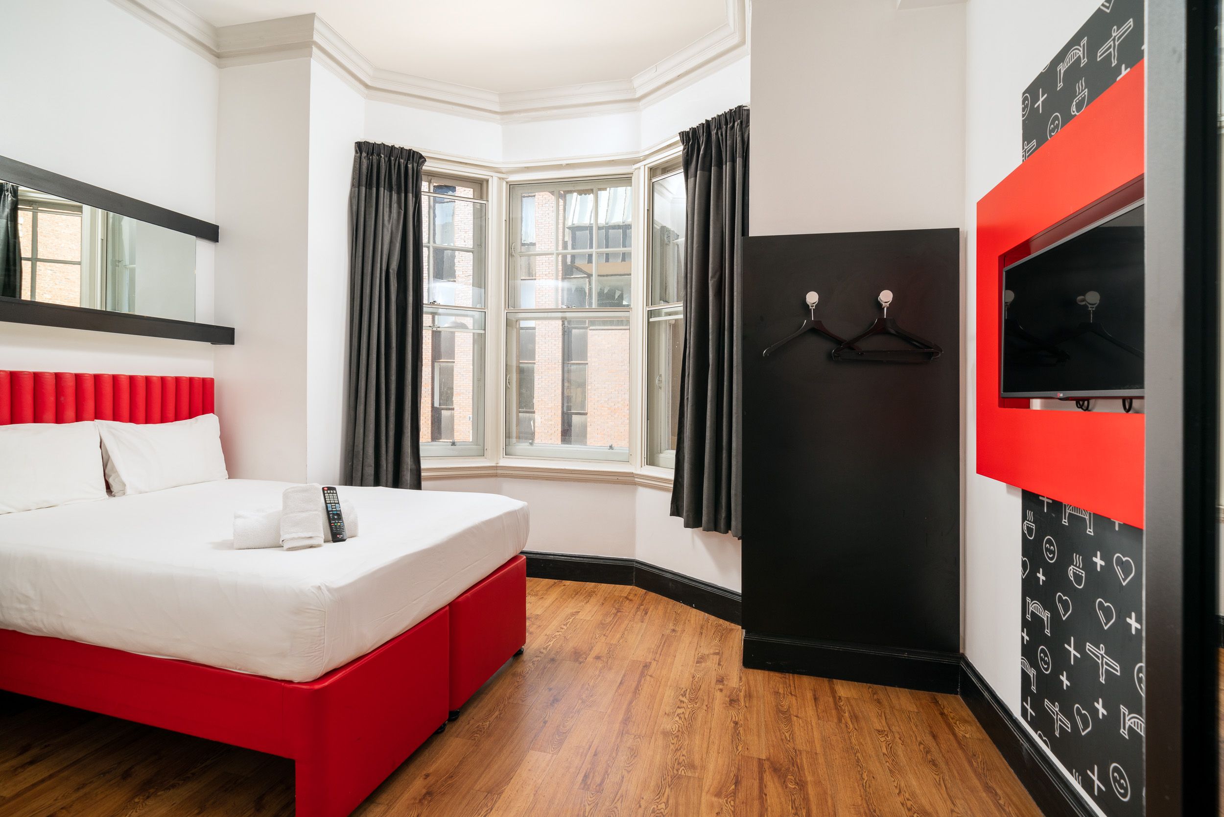 Modern easyHotel room in Newcastle featuring a double bed with red base and headboard, a bright bay window with dark curtains, and a wall-mounted TV on a vibrant red panel.