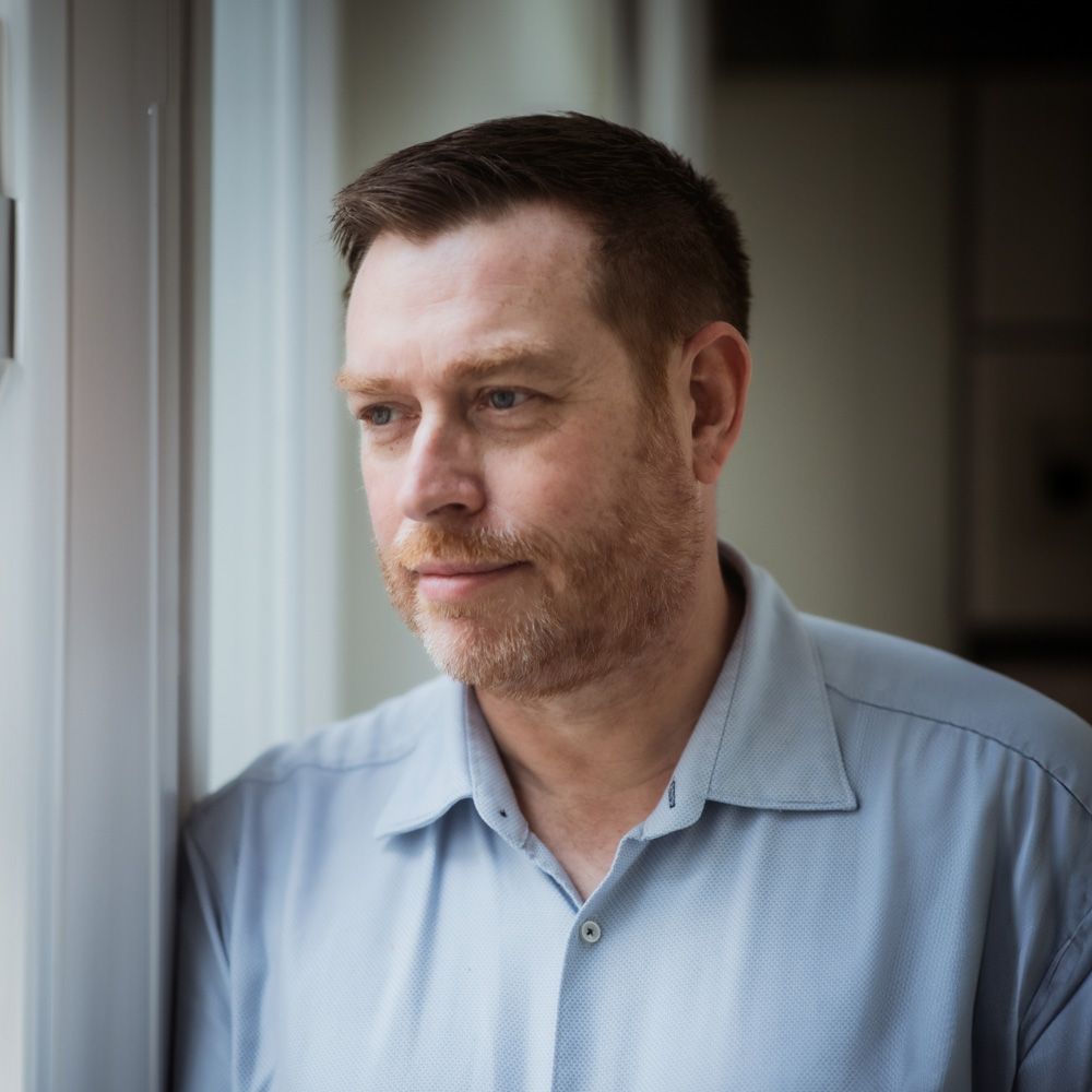 Professional headshot of Nick, a man with a reddish beard and light blue shirt, looking thoughtfully out of a window.