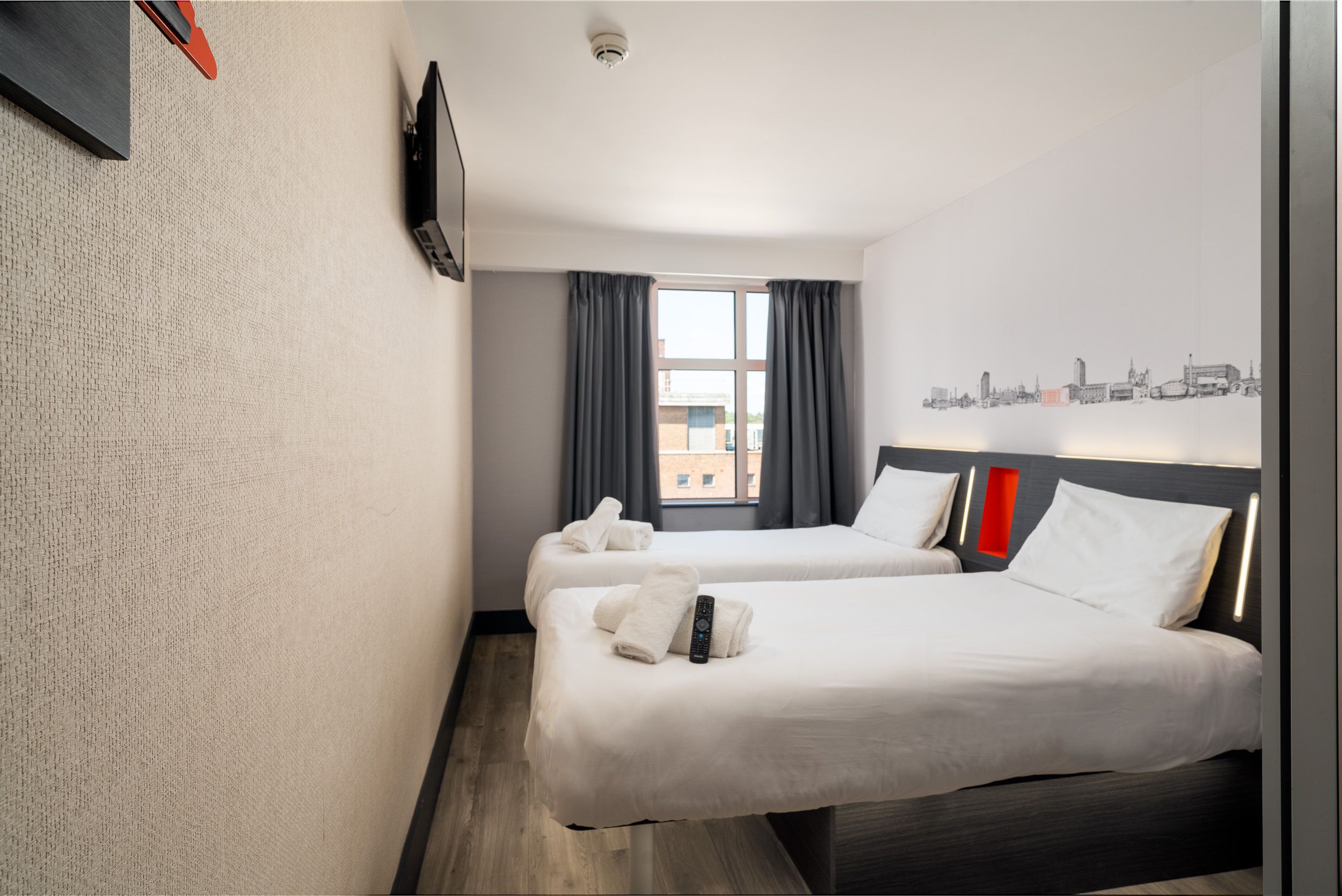 EasyHotel Sheffield twin room with two single beds, crisp white linen, a wall-mounted TV, and a Sheffield city skyline mural above the dark headboards, featuring a large window with grey curtains.