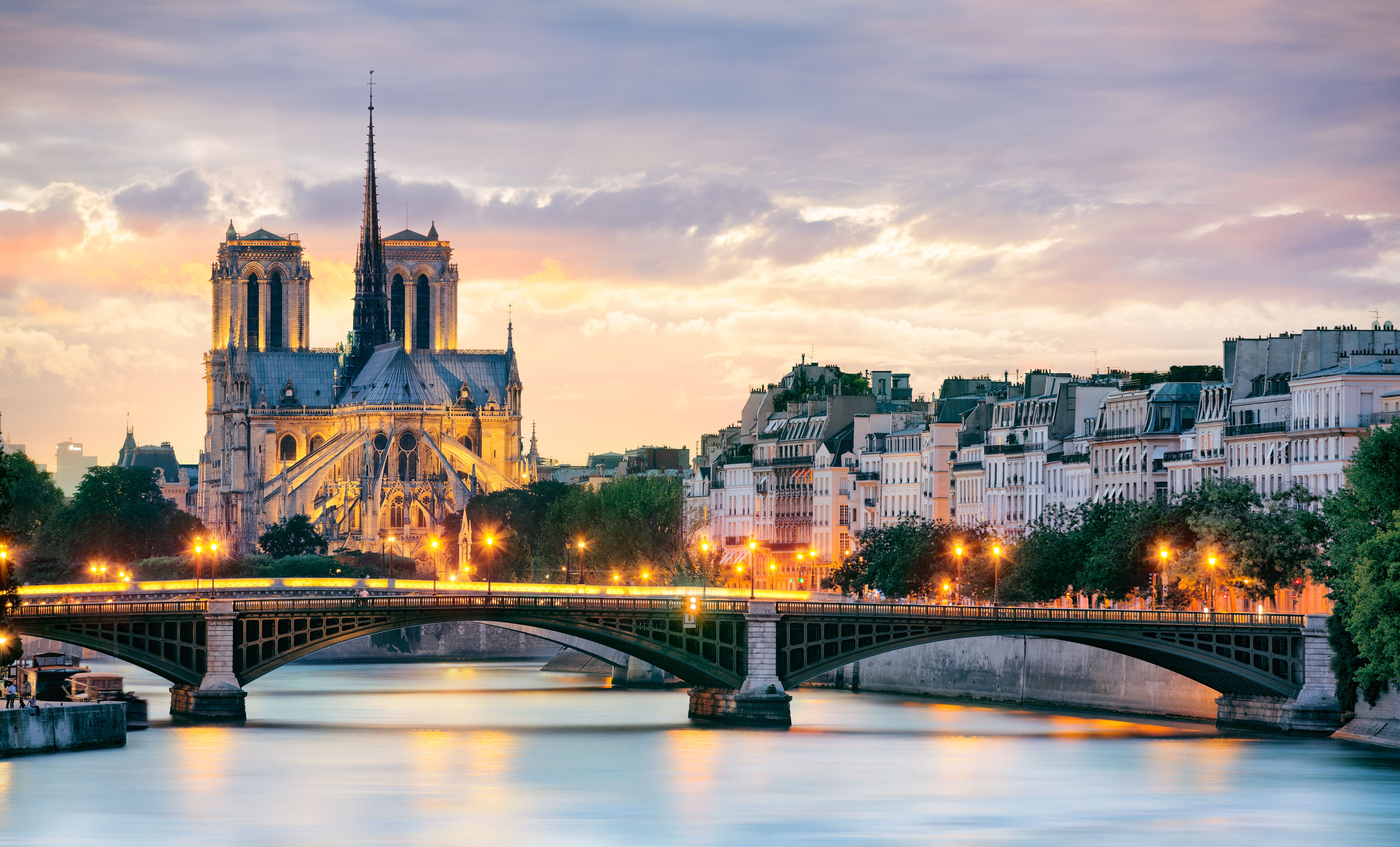 Notre Dame Cathedral and an illuminated bridge over the Seine River at sunset in Paris, France, with classic Parisian buildings along the riverbank.