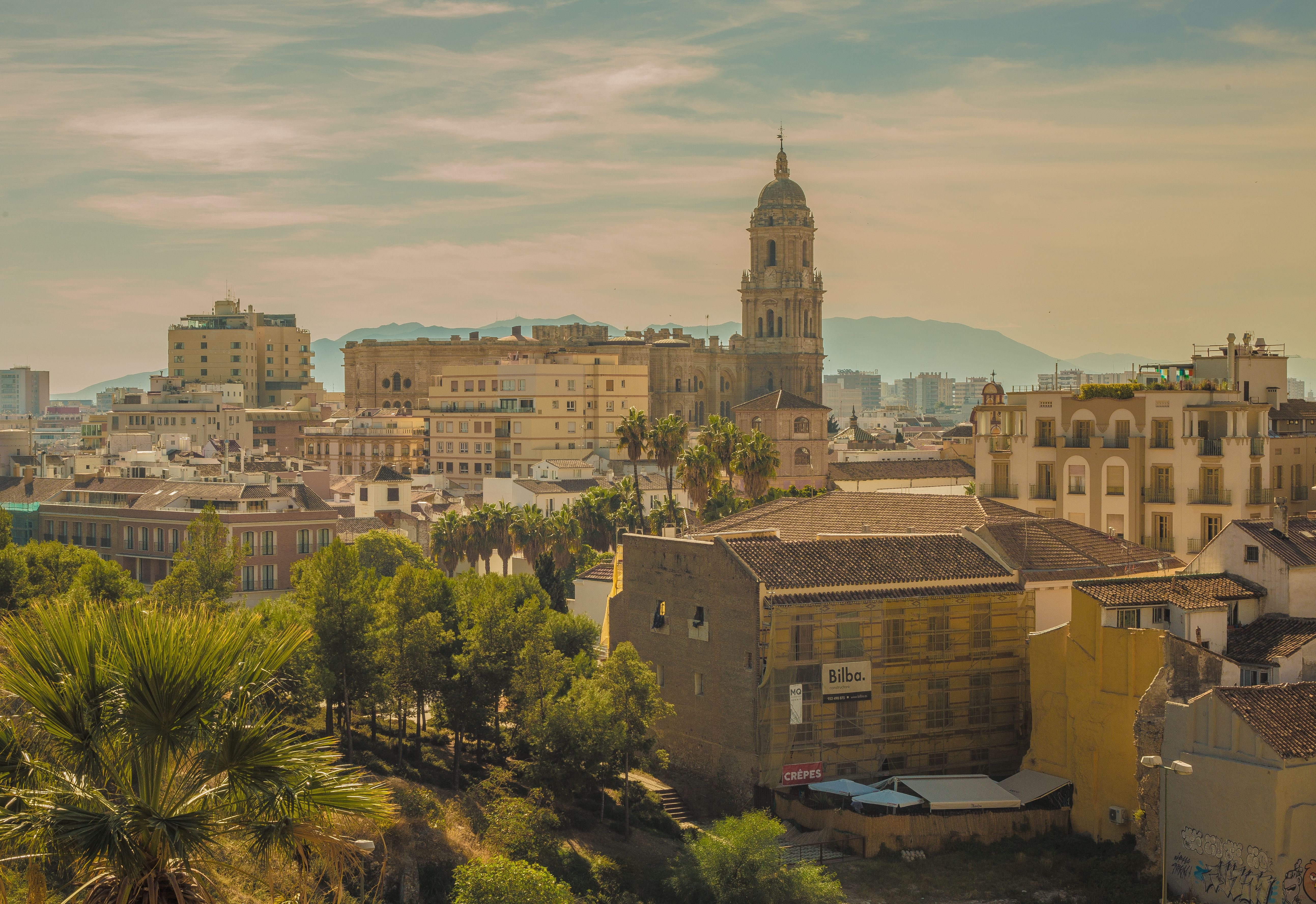 Malaga city skyline featuring the iconic Cathedral tower, historic buildings, lush green trees, and distant mountains under a bright sky.