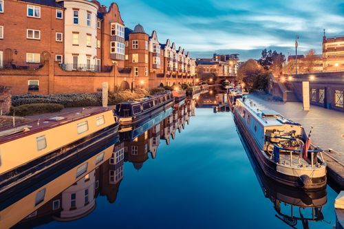 Scenic dusk view of historic Birmingham canals, showcasing traditional narrowboats moored along illuminated brick buildings with reflections on the water.