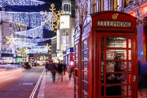 Iconic red London telephone booths on a bustling street at night, brightly lit by elaborate snowflake and icicle Christmas lights overhead. The scene includes blurred people and vehicle light trails.