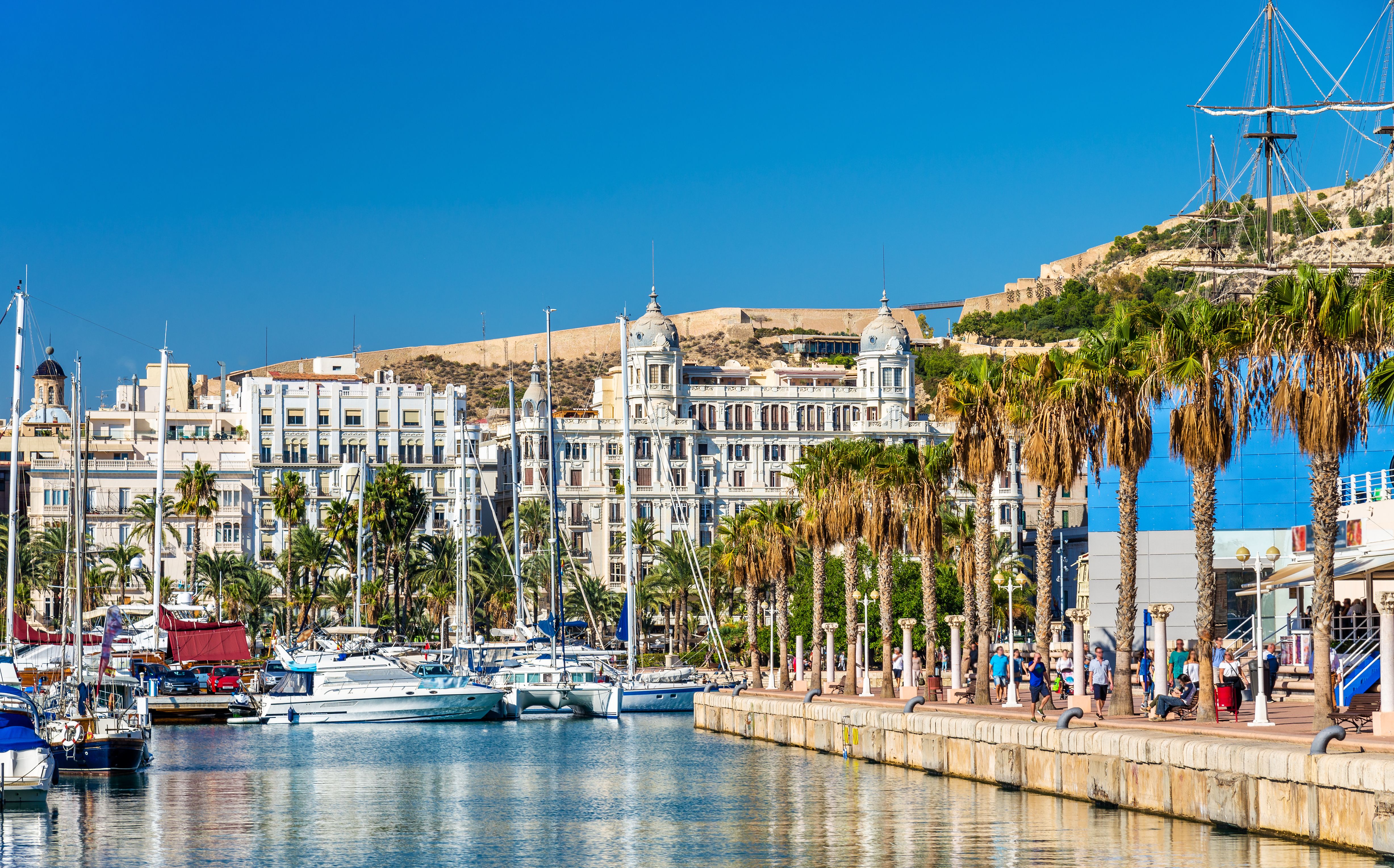 Panoramic view of Alicante marina in Spain, featuring numerous sailboats, a bustling promenade lined with palm trees, ornate white historic buildings including Casa Carbonell, and Santa Bárbara Castle perched on a hill under a clear blue sky.