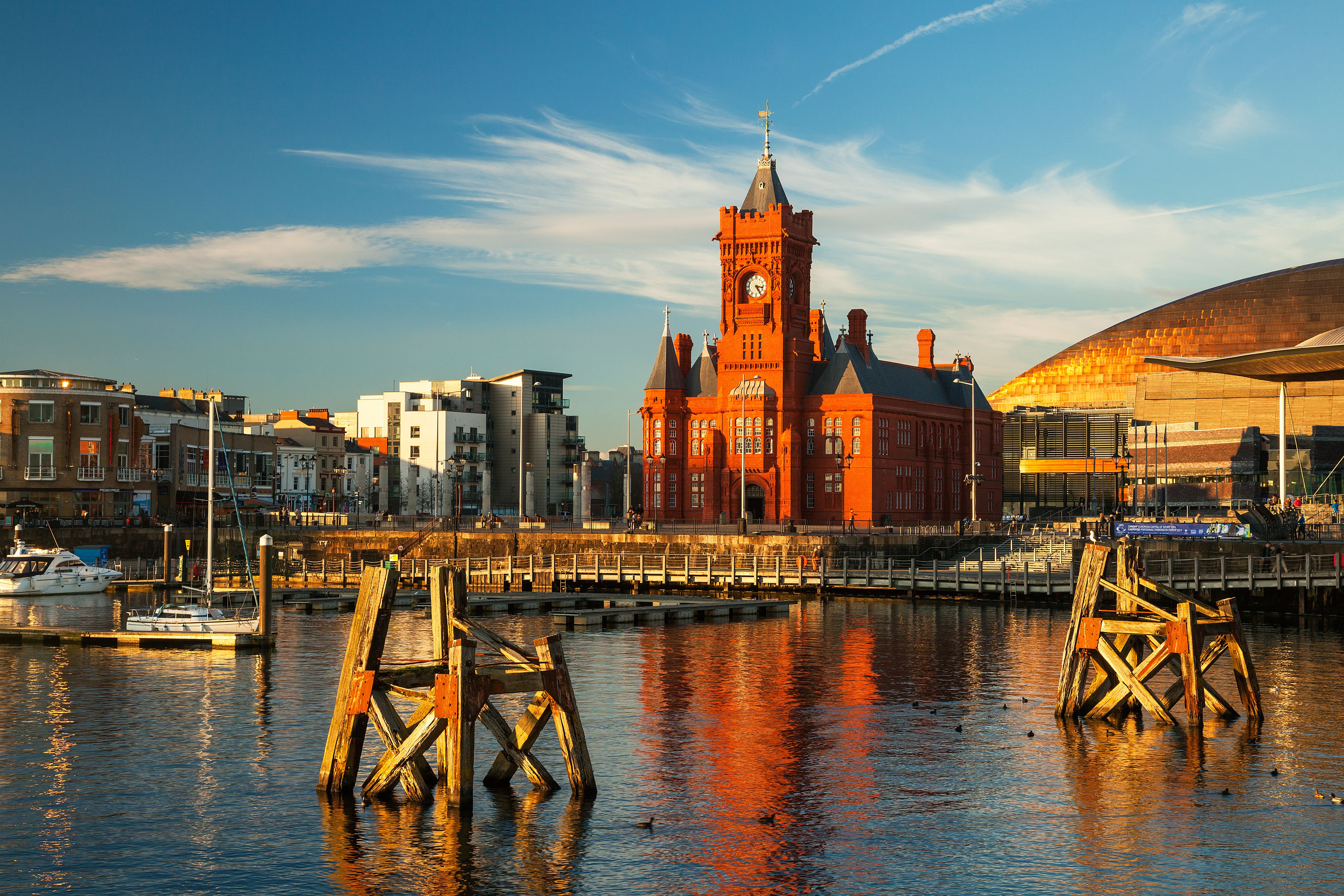 Iconic Welsh architecture in Cardiff Bay: the historic red brick Pierhead Building with a clock tower and the modern Wales Millennium Centre, reflected in the calm bay waters under a bright blue sky.