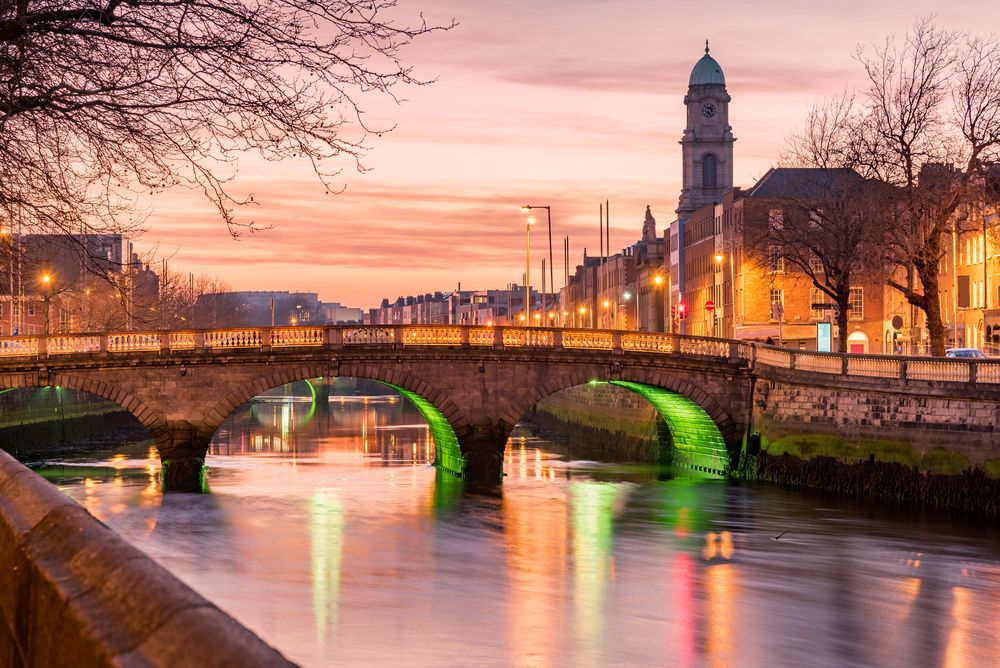 Dublin's O'Connell Bridge over the River Liffey illuminated with vibrant green lights at dusk, reflecting on the water, with city buildings and a prominent clock tower under a colorful sunset sky.