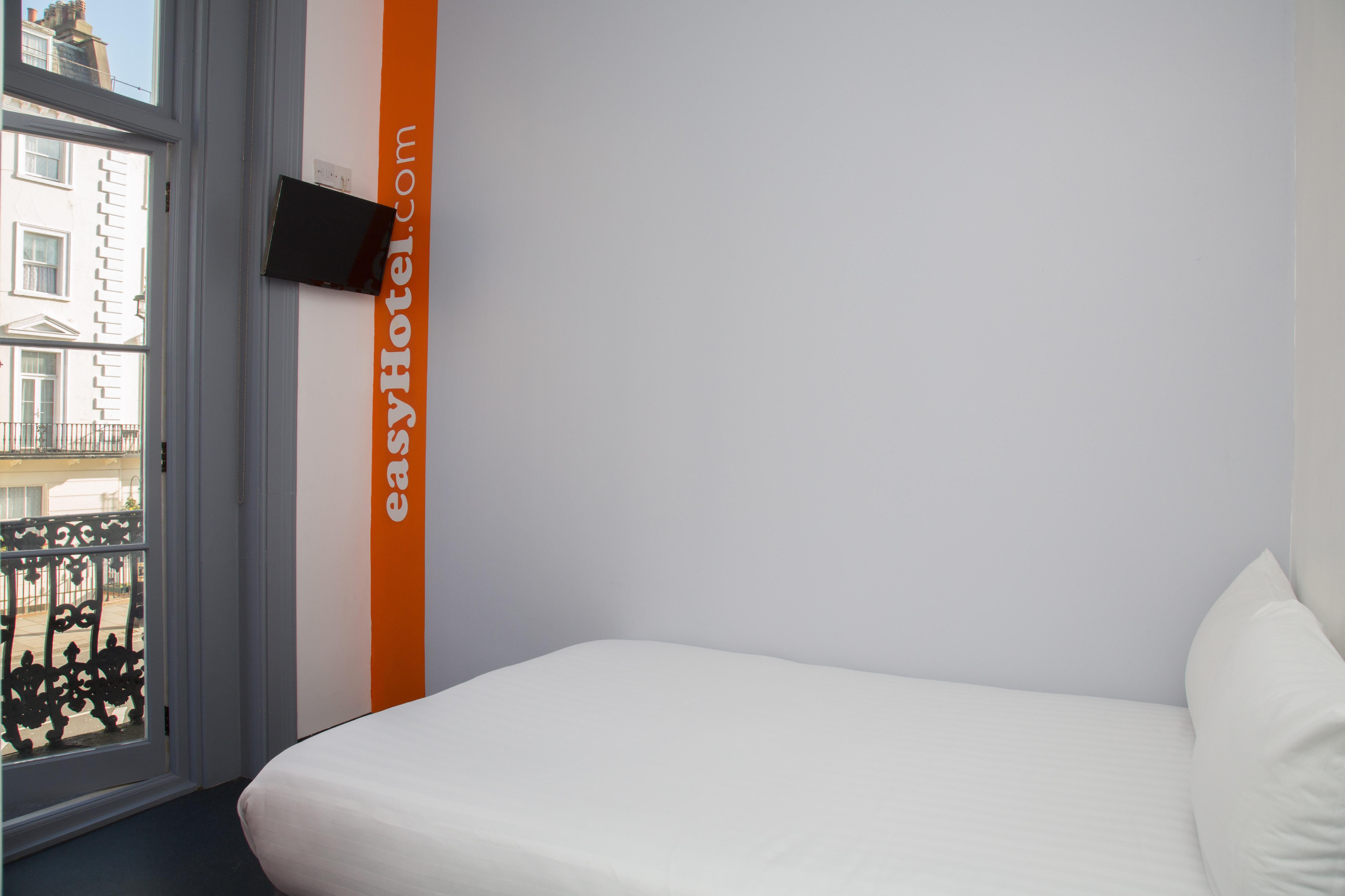 Minimalist easyHotel room in London Victoria with a white bed, wall-mounted TV, and balcony overlooking classic Victorian architecture.